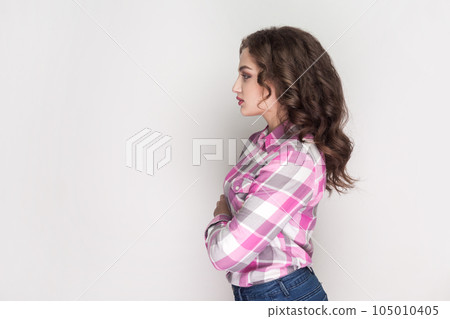 Side view portrait of serious woman with curly hair standing with crossed hands, looking ahead with strict expression, wearing pink checkered shirt. Indoor studio shot isolated on gray background. Side view portrait of serious woman with curly hair standing with crossed hands, looking ahead with strict expression, wearing pink checkered shirt. Indoor studio shot isolated on gray background. 105010405