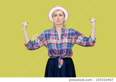 Portrait of mature woman wearing checkered shirt and hat raises hands and shows muscles, feels proud about her achievements in gym, smiles broadly. Indoor studio shot isolated on yellow background. 105010407