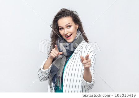 Portrait of overjoyed happy positive brunette woman pointing at camera with index fingers, choosing you, wearing striped jacket and scarf. Indoor studio shot isolated on gray background. Portrait of overjoyed happy positive brunette woman pointing at camera with index fingers, choosing you, wearing striped jacket and scarf. Indoor studio shot isolated on gray background. 105010419
