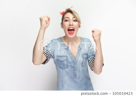 Portrait of extremely happy cheerful blonde woman wearing blue denim shirt and red headband standing clenched fists, celebrating victory. Indoor studio shot isolated on gray background. Portrait of extremely happy cheerful blonde woman wearing blue denim shirt and red headband standing clenched fists, celebrating victory. Indoor studio shot isolated on gray background. 105010423