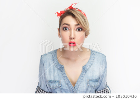 Portrait of astonished surprised blonde woman wearing blue denim shirt and red headband standing looking at camera with big eyes, being amazed. Indoor studio shot isolated on gray background. 105010436