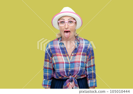 Portrait of childish mature woman wearing checkered shirt, hat and eyeglasses sticking out tongue, making funny grimace. Indoor studio shot isolated on yellow background. 105010444