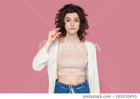 Portrait of confused disappointed beautiful woman with curly hairstyle wearing casual style outfit showing small size with fingers. Indoor studio shot isolated on pink background. Portrait of confused disappointed beautiful woman with curly hairstyle wearing casual style outfit showing small size with fingers. Indoor studio shot isolated on pink background. 105010450