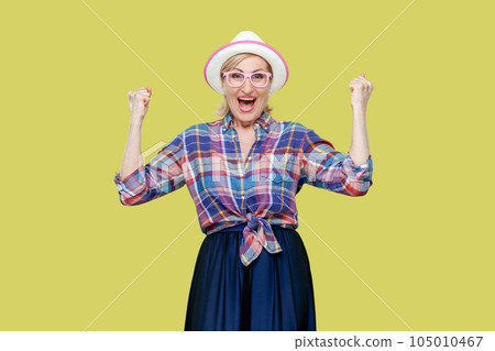 Overjoyed senior woman in checkered shirt, hat, eyeglasses clenches fists with happiness, opens mouth widely as shouts loudly, celebrates her success. Indoor studio shot isolated on yellow background. Overjoyed senior woman in checkered shirt, hat, eyeglasses clenches fists with happiness, opens mouth widely as shouts loudly, celebrates her success. Indoor studio shot isolated on yellow background. 105010467