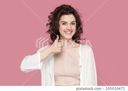 Nice joke. Portrait of woman with curly hairstyle wearing casual style outfit keeps thumb raised, being in good mood, shows her agreement. Indoor studio shot isolated on pink background. Nice joke. Portrait of woman with curly hairstyle wearing casual style outfit keeps thumb raised, being in good mood, shows her agreement. Indoor studio shot isolated on pink background. 105010471