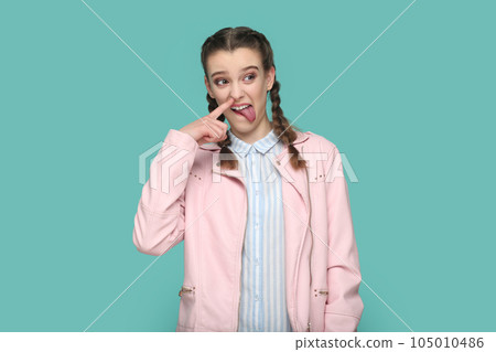 Portrait of funny childish teenager girl with braids wearing pink jacket standing with finger in nose, sticking tongue out, looking away. Indoor studio shot isolated on green background. Portrait of funny childish teenager girl with braids wearing pink jacket standing with finger in nose, sticking tongue out, looking away. Indoor studio shot isolated on green background. 105010486