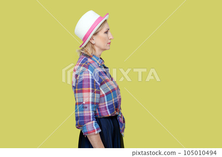 Side view portrait of serious strict mature woman wearing checkered shirt and hat standing looking ahead, being in bad mood. Indoor studio shot isolated on yellow background. 105010494