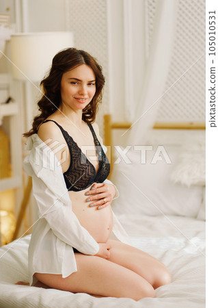 a slender pregnant woman in a black underwea and white shirt on the bed 105010531