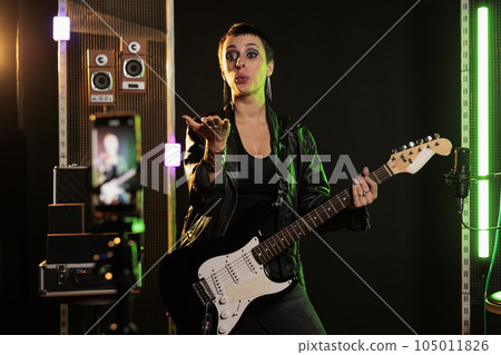 Rockstar blowing air kisses in front of smartphone camera while playing at electric guitar in sound studio, working at rock album using electricinstrument. Rebel musician recording heavy metal song Rockstar blowing air kisses in front of smartphone camera while playing at electric guitar in sound studio, working at rock album using electricinstrument. Rebel musician recording heavy metal song 105011826