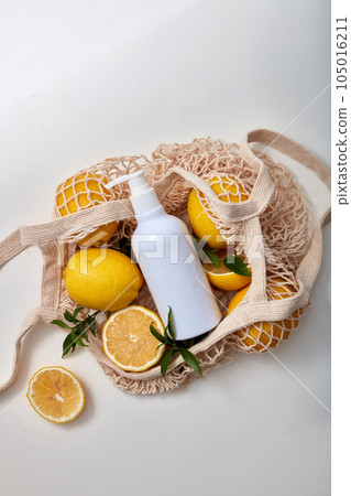 Top view of a white pump bottle unlabeled displayed on a mesh bag full of fresh lemons and green leaves isolated on white background. Minimal scene for advertising cosmetic product of lemon extract 105016211