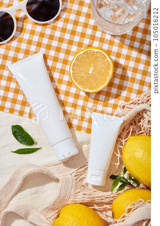 Lifestyle concept for advertising cosmetic with lemon ingredient. Two plastic bottles unbranded decorated with fresh lemon on mesh bag and sunglasses on yellow checkered fabric background 105016212
