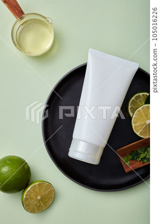 Mockup scene for advertising with white bottle unbranded for cosmetic, facial cleanser or cream of lime extract. Fresh lime and lime slices decorated on round black plate on a pastel background. 105016226