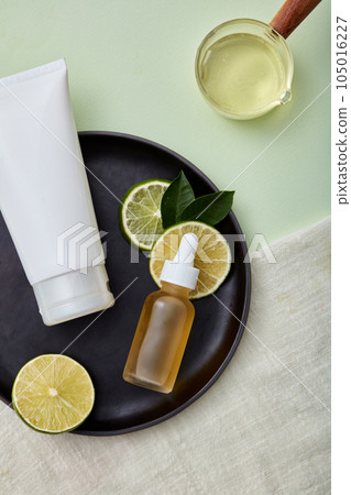 Top view of empty bottles for beauty products, cosmetics with natural origin extracted from lime. Advertising scene with pastel background, fresh lime slices and leaves on black plate and gray towel. 105016227