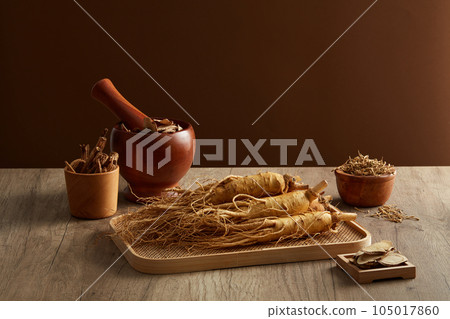 Ginseng roots and medicine herbs on wooden tray decorated with  wooden mortar and pestle on brown background. Herbal for the preparation of a tonic drink. Photography traditional medicine content 105017860