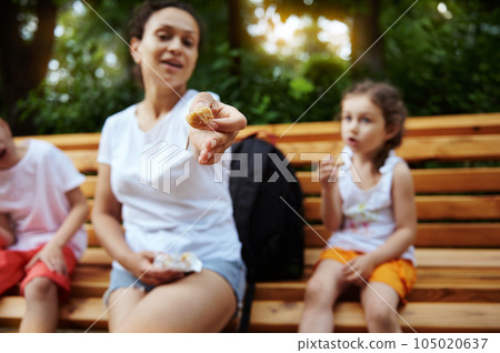 Focus on a dried figs fruit in the hands of a smiling woman, cheerful mother chilling out with her kids in the city park Focus on a dried figs fruit in the hands of a smiling woman, cheerful mother chilling out with her kids in the city park 105020637