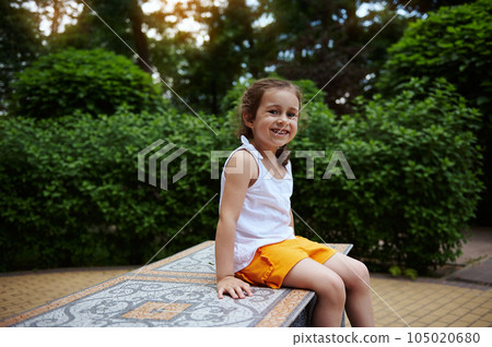 Mischievous little child, preschool girl 4 years smiling looking at camera, sitting on a mosaic bench in the city park Mischievous little child, preschool girl 4 years smiling looking at camera, sitting on a mosaic bench in the city park 105020680