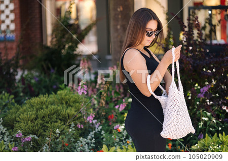 Photo of process when female taking something from a white knitted bag. Young woman in black dress and glasses 105020909