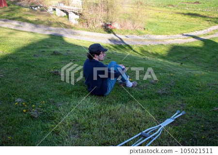 Young man sits alone on grass hill in shadow of a house with crutches 105021511
