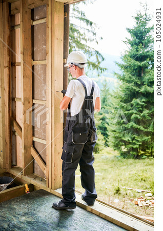 Carpenter constructing wooden framed house. Man worker in glasses working with screwdriver, wearing work overalls and helmet. Concept of modern eco-friendly construction. 105024931