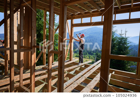 Carpenter constructing wooden frame, two-story house near forest. Bearded man hammering nails with hammer while wearing protective helmet and work coveralls. Concept of modern ecological construction. 105024936