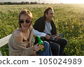 Summer time. Beautiful young couple enjoying time in field around grass. Lifestyle, love, dating, vacation concept. Woman drinking beer, man playing ukulele 105024989