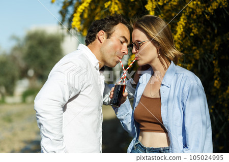 Young happy couple outdoors drinking cold drink from glass bottle with straw. Side view of man and woman drinking soft drink and smiling. 105024995