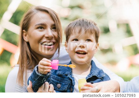Beautiful cute young mom with her cute little son having fun sitting on the grass in the park outdoors and blowing soap bubbles Beautiful cute young mom with her cute little son having fun sitting on the grass in the park outdoors and blowing soap bubbles 105027093
