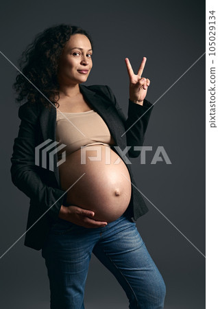 Attractive young pregnant woman posing bare belly on gray studio background, smiling and showing peace sign with fingers 105029134