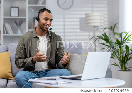 Young Latin American businessman, freelancer works remotely from home. Sitting on the sofa in a headset and talking on a video call through a laptop. 105029689
