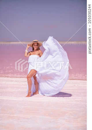 Woman in pink salt lake. She in a white dress and hat enjoys the scenic view of a pink salt lake as she walks along the white, salty shore, creating a lasting memory. 105030244