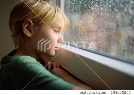 Portrait of a sad preteen boy standing at the window. Child watching the rain outside. Post-traumatic disorder. Accommodation of grief. Experiencing loss. Fear. Depression. Stress. 105030265