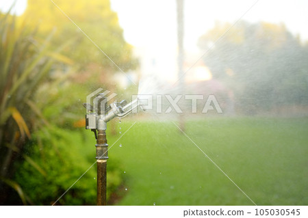 Garden sprinkler in a public park. Gardening equipment. Watering the grass in the yard on a sunny summer day Garden sprinkler in a public park. Gardening equipment. Watering the grass in the yard on a sunny summer day 105030545