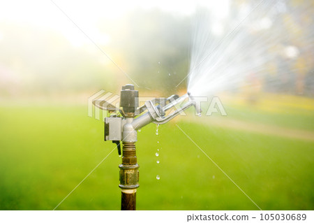 Garden sprinkler in a public park. Gardening equipment. Watering the grass in the yard on a sunny summer day Garden sprinkler in a public park. Gardening equipment. Watering the grass in the yard on a sunny summer day 105030689