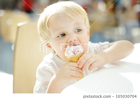 Cute little boy eating ice cream in a waffle cone. Child is tasting gelato in Italian cafe. Unhealthy street fast food. 105030704