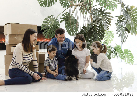 Happy family sitting on the floor in living room. Funny kids with little dog by carton boxes, parents laughing Happy family sitting on the floor in living room. Funny kids with little dog by carton boxes, parents laughing 105031085