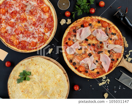 Three different pizzas on a black background with cooking ingredients. Top view, pizzeria. Three different pizzas on a black background with cooking ingredients. Top view, pizzeria. 105031147