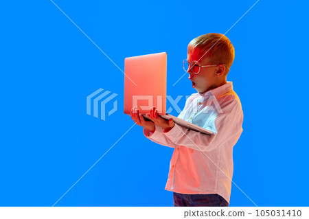Online lessons. Emotional boy, child in glasses looking with shocked face in laptop against blue studio background in neon light 105031410