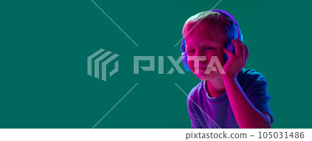 Child, little boy listening to music in headphones against cyan studio background in neon light. Grimacing face 105031486