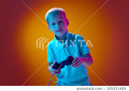 Playful little boy, child playing console against gradient yellow studio background in neon light. Leisure time and games 105031489