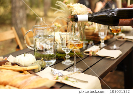Glass of orange wine placed near dishware and bouquet of fresh flowers amidst assorted fruits on wooden table 105031522