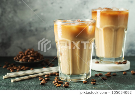Ice coffee in a tall glass with cream poured over, ice cubes and beans on a dark concrete table. 105034092