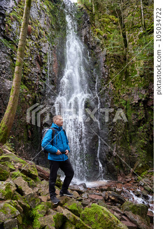 Tourist attraction of Germany - falls of Burgbach Waterfall near Schapbach, Black Forest, Baden-Wurttemberg, Germany. Man hiker in blue jacket standing on stone and looks at flow of falling water Tourist attraction of Germany - falls of Burgbach Waterfall near Schapbach, Black Forest, Baden-Wurttemberg, Germany. Man hiker in blue jacket standing on stone and looks at flow of falling water 105034722