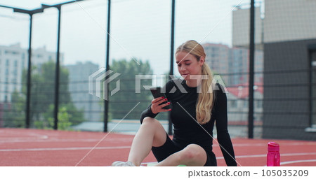 Young woman on a basketball court using smartphone. Girl resting after playing basketball, reading messages. Young woman on a basketball court using smartphone. Girl resting after playing basketball, reading messages. 105035209