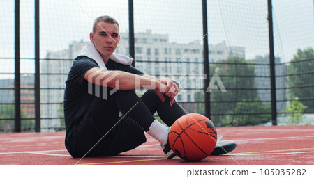 Male player sitting on basketball sports ground with ball. Concentrated sportsman looking at camera during training. Male player sitting on basketball sports ground with ball. Concentrated sportsman looking at camera during training. 105035282