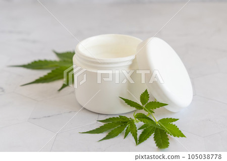 Opened white cream jar with a lid near cannabis leaves close up. Cosmetic Mockup Opened white cream jar with a lid near cannabis leaves close up. Cosmetic Mockup 105038778