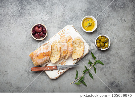 Sliced fresh ciabatta on cutting kitchen board, green and brown olives, olive oil with rosemary, olive tree branches on gray concrete stone rustic background top view copy space 105039970