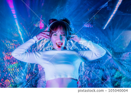 Portrait of emotional screaming woman in white clothes and headphones dancing in neon light. Dj , music lover. White dress code party. Silent disco. Enjoy moment on shiny background. Selective focus. 105040004