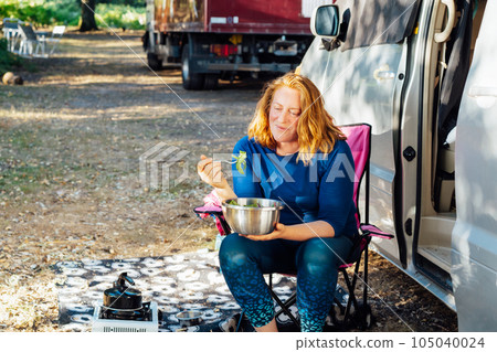 Relaxed Middle aged woman eating veggie salad outside camper van in the forest camp during road trip lunch. Enjoying nomad lifestyle, active weekend, vacation. 105040024