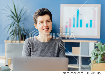 Portrait of smiling neutral gender middle aged woman at office desk with laptop and chart board with financial graphs on background. Business analysis concept. Data reporting. financial performance Portrait of smiling neutral gender middle aged woman at office desk with laptop and chart board with financial graphs on background. Business analysis concept. Data reporting. financial performance 105040055