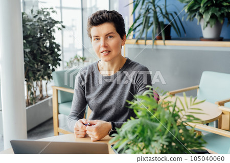 Neutral gender middle aged woman with no make-up in casual clothing using laptop and smiling while working indoors in her workstation in an open space office. Online video call, IT HR, recruiting 105040060